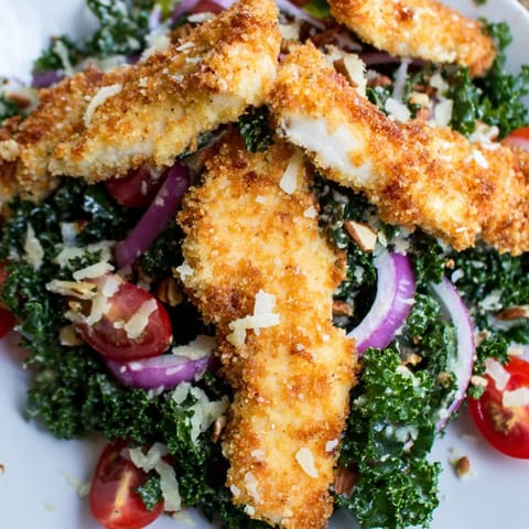 Golden, crispy chicken strips rest atop a vibrant kale salad, drizzled with tangy maple-Dijon dressing and topped with halved cherry tomatoes.