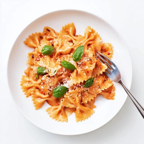 A close-up of silky tomato sauce coating farfalle pasta, highlighted by chopped basil and a sprinkle of cheese.