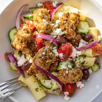 Crispy Chicken Greek Pasta features golden fried chicken bites, al dente short pasta, and a vibrant salad of tomatoes, cucumber, feta, and olives.